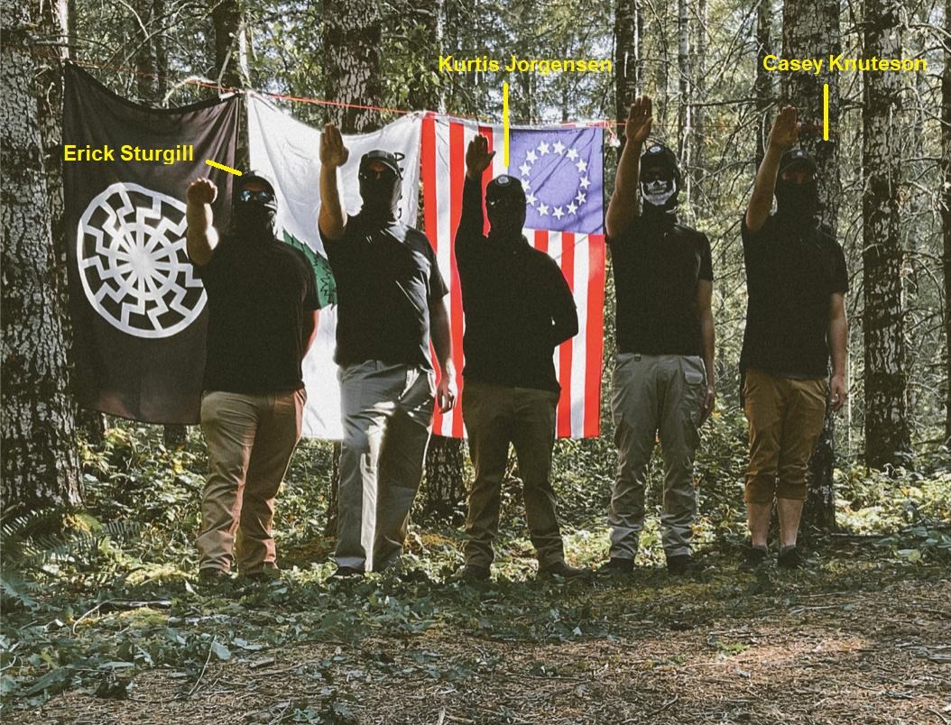 Picture of five RCN members standing in Tillamook forest; they are throwing Nazi salutes in front of fascist flags; Erick, Kurtis Jorgensen, and Casey Knuteson are labeled